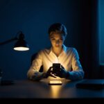 Uma figura está sentada em uma mesa mal iluminada, digitando em um smartphone com uma expressão secreta. O quarto está escuro, com apenas o brilho do telefone iluminando seu rosto.